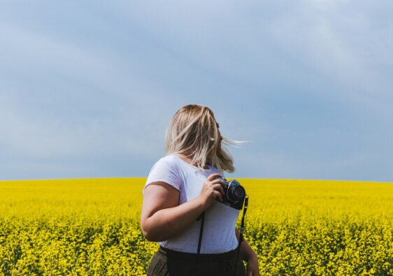A fat woman with a camera in front of a rapeseed field and a blue sky.