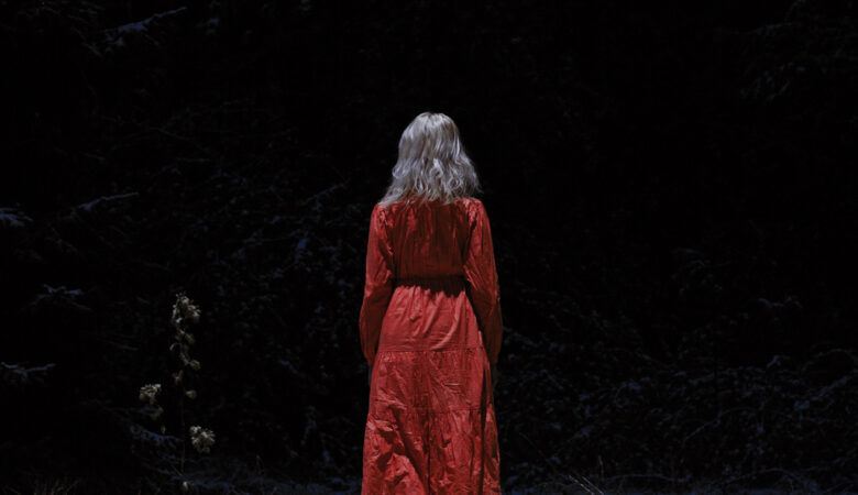 The back of a blond woman in a red dress, facing the darkness.